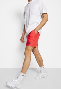 Muž v bílém tričku, červených šortkách Nike, bílých ponožkách Nike a bílých teniskách, kráčející proti jednoduchému bílému pozadí.