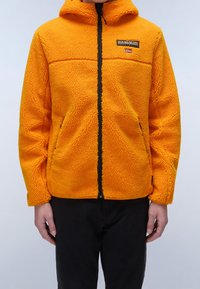 Giacca in pile di un vivace arancione con superficie testurizzata, chiusura frontale con zip e cappuccio. Presenta una zip nera e un'etichetta sul petto con un emblema.