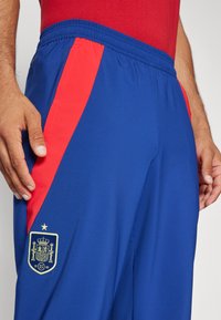 Pantalones deportivos azules con rayas laterales rojas, cinturilla elástica y un emblema nacional bordado en dorado en el muslo izquierdo.