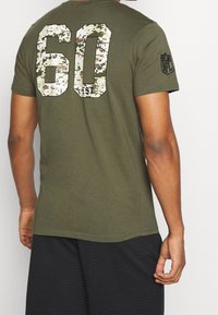 Olivgrön t-shirt med stort, pixelerat kamouflage "60" och "EST" tryckt på ryggen. Kort ärm, tillverkad av mjukt bomullsmaterial.