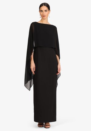 Frau trägt ein bodenlanges schwarzes Kleid mit transparenten, fließenden Umhangärmeln, steht aufrecht, Haare zurückgebunden und trägt Perlenohrringe.