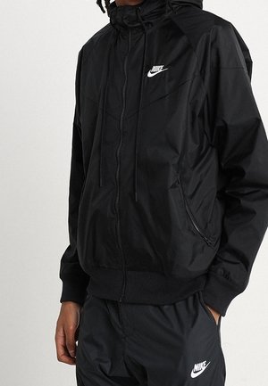 Personne portant une veste coupe-vent noire Nike avec fermeture éclair et un pantalon Nike noir assorti avec des logos sur la poitrine et la cuisse.