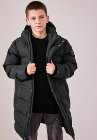Manteau en doudoune noir avec capuche, doté d'une fermeture éclair à l'avant et de boutons-pression, design matelassé et poignets élastiques, porté sur un pull noir.