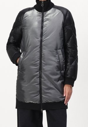 Langer gesteppter Mantel mit glänzend silberner Vorderseite und schwarzen Ärmeln, Reißverschluss, Seitentaschen und hohem Kragen, getragen mit schwarzer Hose.