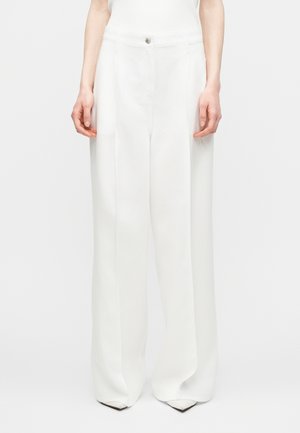 Donna che indossa pantaloni bianchi a vita alta e gamba larga con piega frontale e chiusura con bottone, abbinati a scarpe bianche a punta.