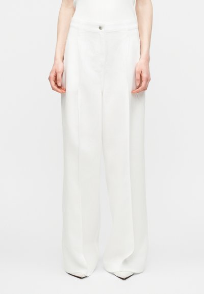 Femme portant un pantalon blanc à taille haute, jambes larges, avec pli frontal et fermeture à boutons, associé à des chaussures blanches à bout pointu.