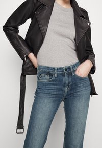 Person som bär blå jeans med händerna i fickorna, en ljusgrå t-shirt och en svart läderjacka med ett oöppnat bälte som hänger ner.