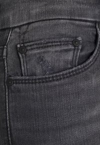Los jeans de mezclilla gris presentan un bolsillo frontal con dos botones negros y costura visible. La tela muestra una textura fina y un ligero desvanecimiento.