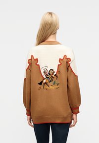 Γυναίκα που φοράει ένα καφέ και κρεμ πουλόβερ με πολύχρωμο κεντητό σχέδιο ενός καουμπόη που καβαλάει άλογο στην πλάτη, συνδυασμένο με σκούρο παντελόνι.