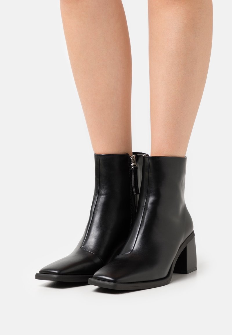 Bottines en cuir noir avec un bout pointu, un talon carré et une fermeture éclair sur le côté. Texture lisse et design minimaliste.