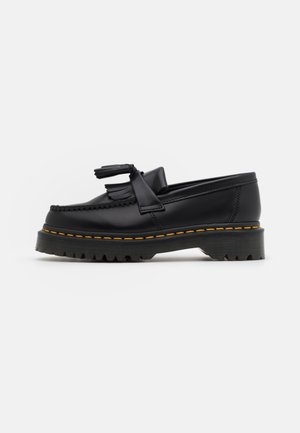 Zwarte leren loafer met kwastjes, franjes, dikke geribbelde zool en gele stiksels langs de rand van de zool, zijaanzicht.