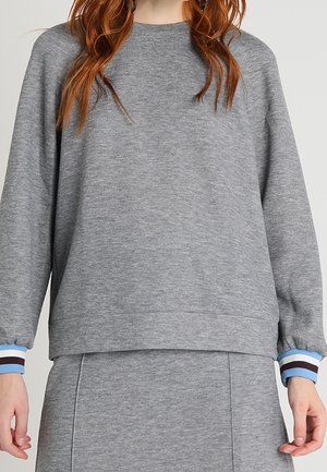 Kvinde iført en grå langærmet sweatshirt og matchende nederdel, med blå og hvide stribede manchetter og synligt rødt hår.