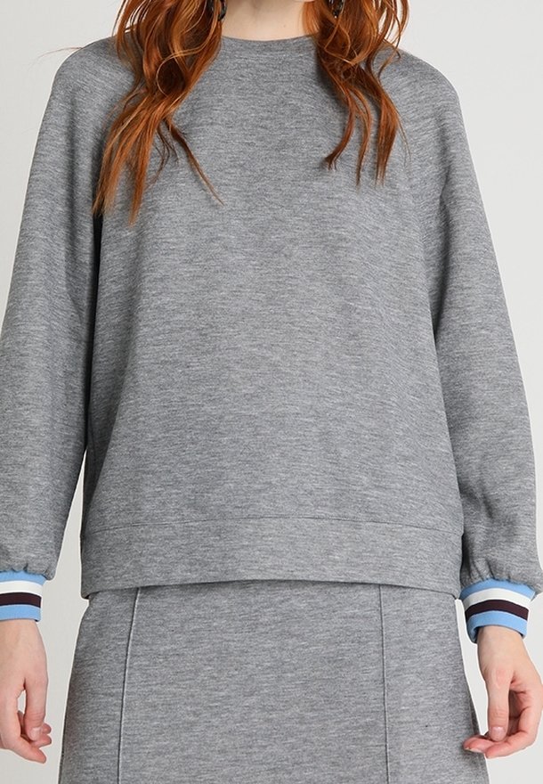 Femme portant un sweat-shirt gris à manches longues et une jupe assortie, avec des poignets rayés bleu et blanc et des cheveux roux visibles.