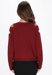 Burgundowy sweter z wycięciami na ramionach i akcentami z koralików. Miękka tekstura, luźny krój i prążkowane wykończenie. Odpowiedni na codzienne wyjścia.