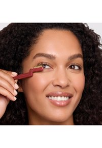 Strumento per ciglia rosso con un'estremità pettinata, tenuto vicino all'occhio, dotato di un manico in plastica e punte metalliche, utilizzato per la cura delle ciglia.