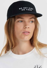 Zwarte pet met een platte rand en geborduurde witte tekst "WE GET HIGH ON TIDES." G draagt een witte T-shirt met ronde hals. Soepele stoftextuur.
