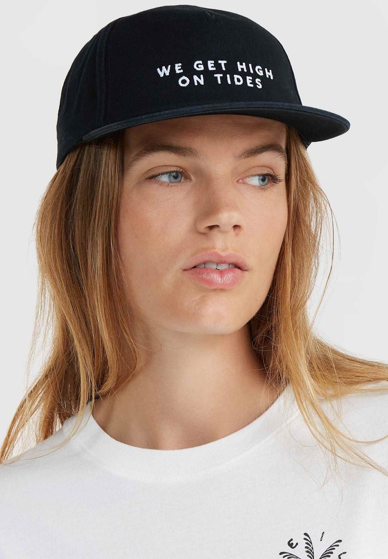 Zwarte pet met een platte rand en geborduurde witte tekst "WE GET HIGH ON TIDES." G draagt een witte T-shirt met ronde hals. Soepele stoftextuur.