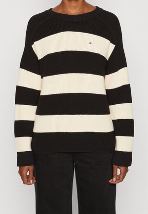 Person wearing black and white wide horizontal striped knit sweater with small logo on chest and black pants, arms relaxed by sides.