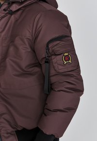 Braune Pufferjacke mit einer Reißverschlusstasche am Ärmel, die ein Logopatch aufweist, elastischen Bündchen und einer Kordelzugkapuze. Glatte, gepolsterte Oberfläche.