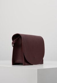 Borsa a mano in pelle bordeaux con superficie testurizzata, chiusura a pattina arrotondata e manico laterale, caratterizzata da un design minimalista e una forma compatta.