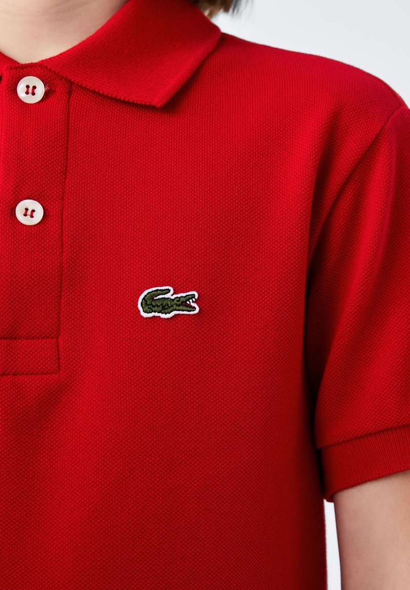 Gros plan d’un polo rouge à manches courtes avec deux boutons blancs et un petit logo crocodile vert sur la poitrine.