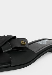 Μαύρο επίπεδο σανδάλι με φαρδιά λουράκι φιόγκο και μικρή χρυσή καρδιά στο λουράκι, μαλακό πέλμα με ορατή επωνυμία.