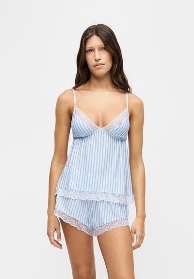 Jeune femme portant un caraco rayé bleu et blanc et un short assorti avec une bordure en dentelle blanche, debout devant un fond clair.