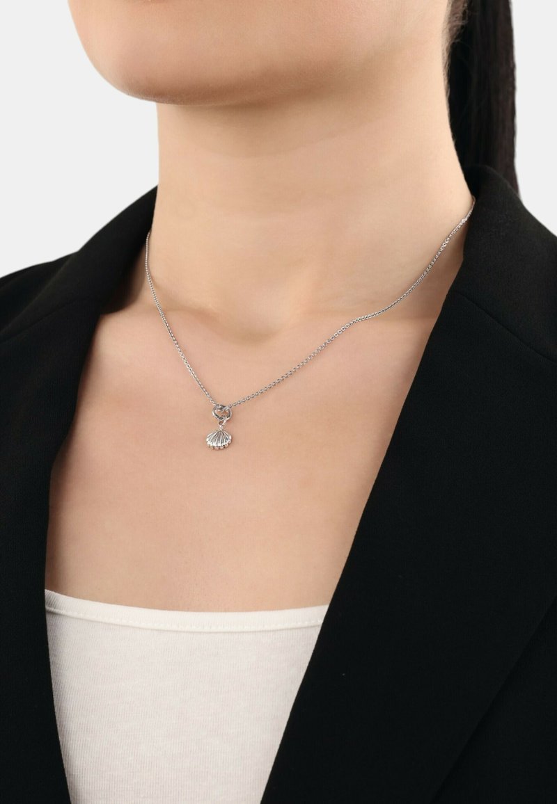 Collana d'argento con ciondolo a forma di conchiglia, attaccata a una delicata catena, indossata sopra una maglietta bianca con un blazer nero.