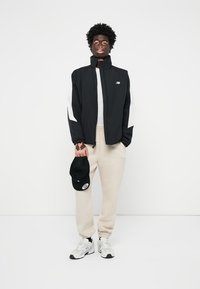 Giacca nera con maniche bianche, pantaloni felpati beige e cappellino nero. Caratterizzata da colletto arrotondato e chiusura con zip. Scarpe sportive completano il look.