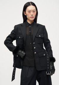 Musta räätälöity takki, jossa on kaksi rintataskua, nepparit ja turkissomisteiset hihat. Vaatteena ohut musta paita ja löysät mustat housut.