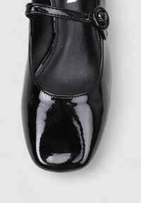 Chaussure Mary Jane en cuir verni noir avec un bout arrondi, une bride de cheville dotée d'une boucle circulaire, et une surface lisse et brillante.
