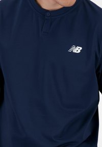 Navy blue long-sleeve shirt with a round neckline, featuring a button detail and a white New Balance logo on the chest, made from smooth fabric.