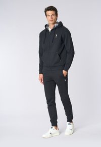 Felpa nera con zip e fodera a quadri, abbinata a pantaloni jogger neri e sneakers bianche. Con logo sul petto.