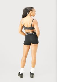 Donna atletica con capelli intrecciati in una coda di cavallo, che indossa un reggiseno sportivo e pantaloni corti neri, calze bianche e scarpe da ginnastica nere, in piedi con le mani sui fianchi.