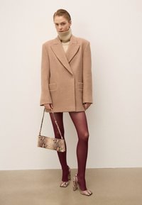 Donna che indossa un cappotto oversize di lana color cammello, un dolcevita beige, collant bordeaux, tacchi in stampa serpente e una borsa a spalla in coordinato con stampa serpente.