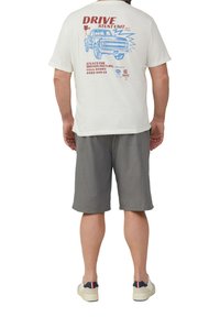 T-shirt blanche avec un imprimé graphique rouge et bleu d'un camion. Un short gris et des baskets de couleur claire complètent la tenue.