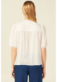 Blusa bianca leggera con maniche corte a sbuffo, delicata rifinitura in pizzo sui polsini e una vestibilità leggermente rilassata con parte posteriore arricciata.