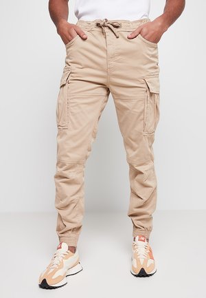 Beige Cargo-Hosen mit mehreren Seitentaschen, elastischen Bündchen und einer verstellbaren Taille. Der Stoff wirkt strapazierfähig und hat eine weiche Textur.