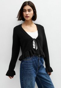 Schwarze Strickjacke mit Rüsche am Saum und den Ärmeln, mit V-Ausschnitt und Bindebandverschluss, getragen über einem weißen Oberteil und Jeans.