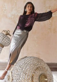 Schwarze Bluse mit Federdesign und durchsichtigen Ärmeln, kombiniert mit einem metallisch silbernen Wickelrock mit seitlichem Schlitz und kreisförmigem Hardware-Detail.