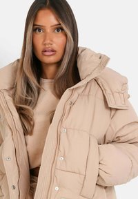 Beige puffjacka med hög krage, tryckknappar och en quiltad textur. Har överdimensionerade axlar och en avslappnad passform.