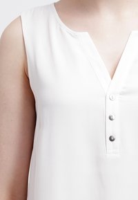 Bezramenná bílá blůza s výstřihem do V, jemnou texturou a třemi stříbrnými knoflíky podél zapínání. Hladký povrch látky.