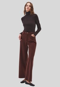 Vrouw die met haar handen in de brede zakken van een bruine broek staat, gekleed in een aangesloten donkerbruine coltrui en zwarte ballerina's, tegen een grijze achtergrond.