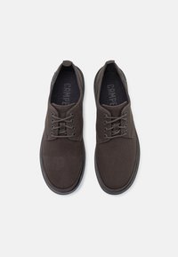 Scarpe in tela marrone con punta tondeggiante e suola piatta in gomma. Presentano lacci e un piccolo etichetta con il logo sul lato. Superficie in tessuto testurizzato.