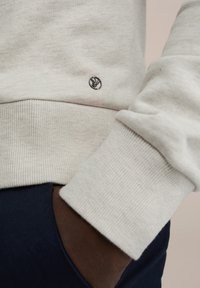 Sweat-shirt gris clair avec poignets côtelés, arborant un petit logo noir. Tissu doux et texturé avec une coupe décontractée, porté sur des vêtements foncés.