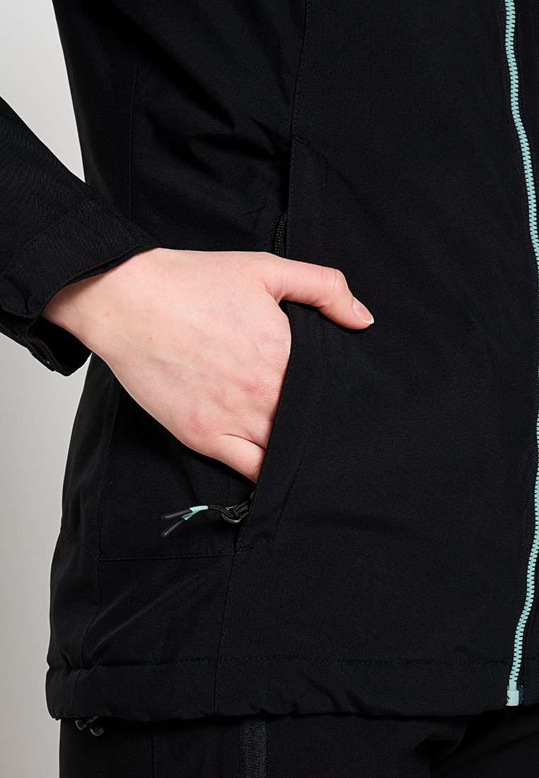 Veste noire avec fermeture éclair sur le devant, une poche latérale zippée et des accents bleu clair le long de la fermeture éclair. Texture lisse et mates.