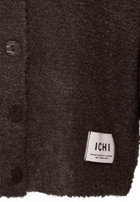 Vêtement texturé marron foncé avec trois boutons et une étiquette blanche indiquant "ICHI" ainsi qu'un petit slogan près du bord inférieur.