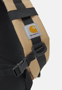 Correia de tecido bege com fita preta e fivela ajustável, apresentando um destacado remendo do logótipo Carhartt em amarelo e cinza.