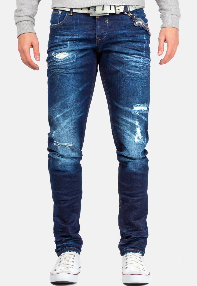 Jeans in denim blu scuro con dettagli abrasivi, inclusi strappi, leggera sbiaditura e una vestibilità slim. Abbinati a sneakers bianche.
