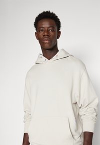 Helle beige Kapuzenjacke aus weichem Baumwollmaterial, mit einer Fronttasche und einem lockeren Schnitt, verstellbarer Kapuze und dezenten Knöpfen.